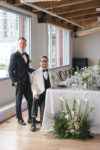 Editorial wedding photography featuring one groom sitting at their reception table and the other groom standing next to him at their luxury Seattle Wedding reception at the Point at Pier 70