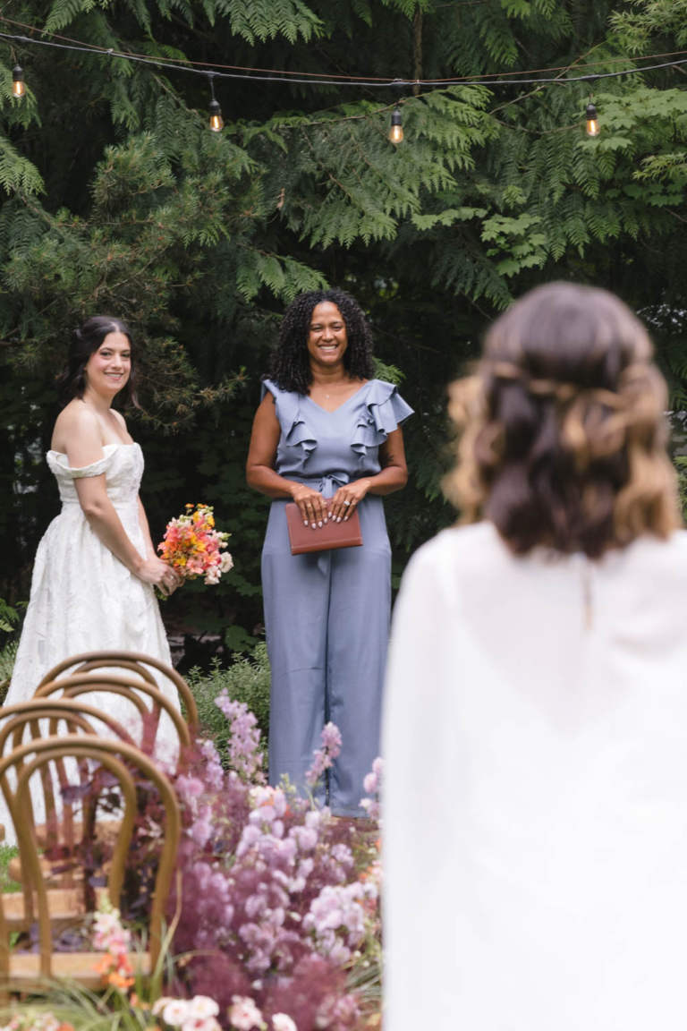 A bride watches her future wife as she walks down the aisle with their smiling officiant in the background.